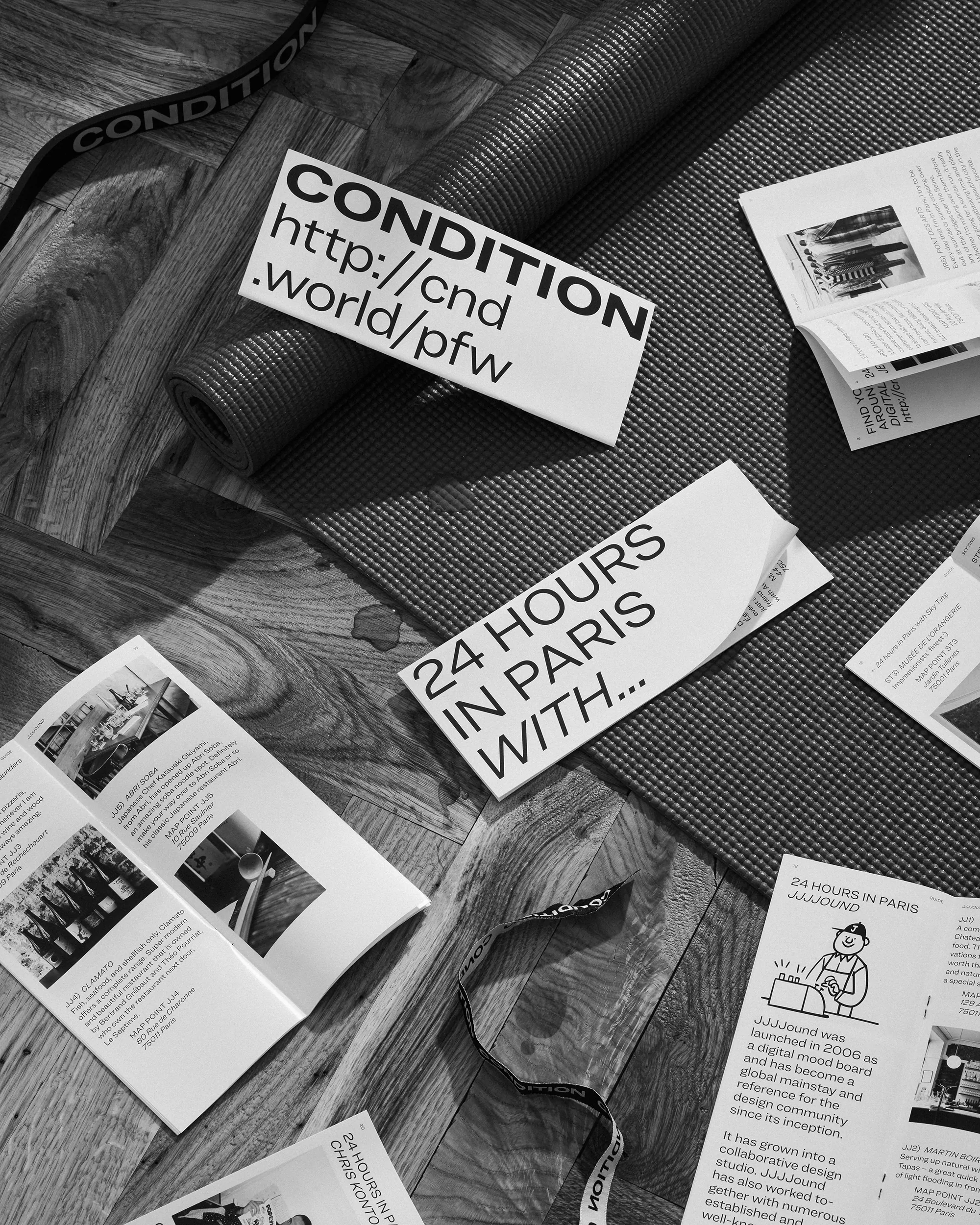 Black and white studio shot of a yoga mat, marketing pamphlets, and a branded strap against a wooden floor surface.