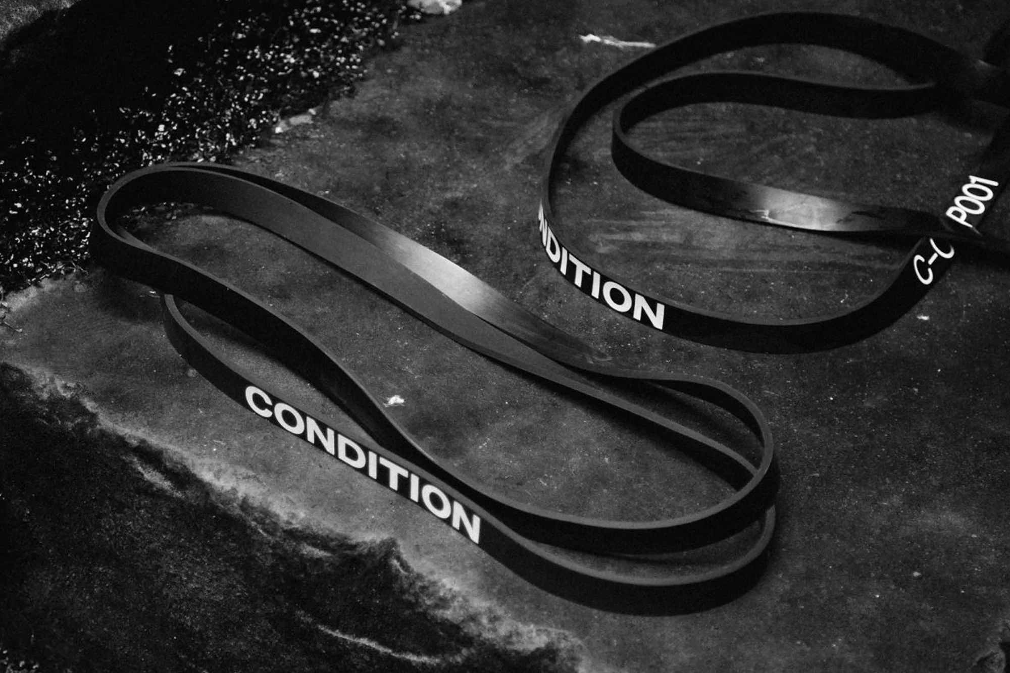 Two black resistance bands lay on a textured dark gray surface, highlighted in a high-contrast black and white photograph.