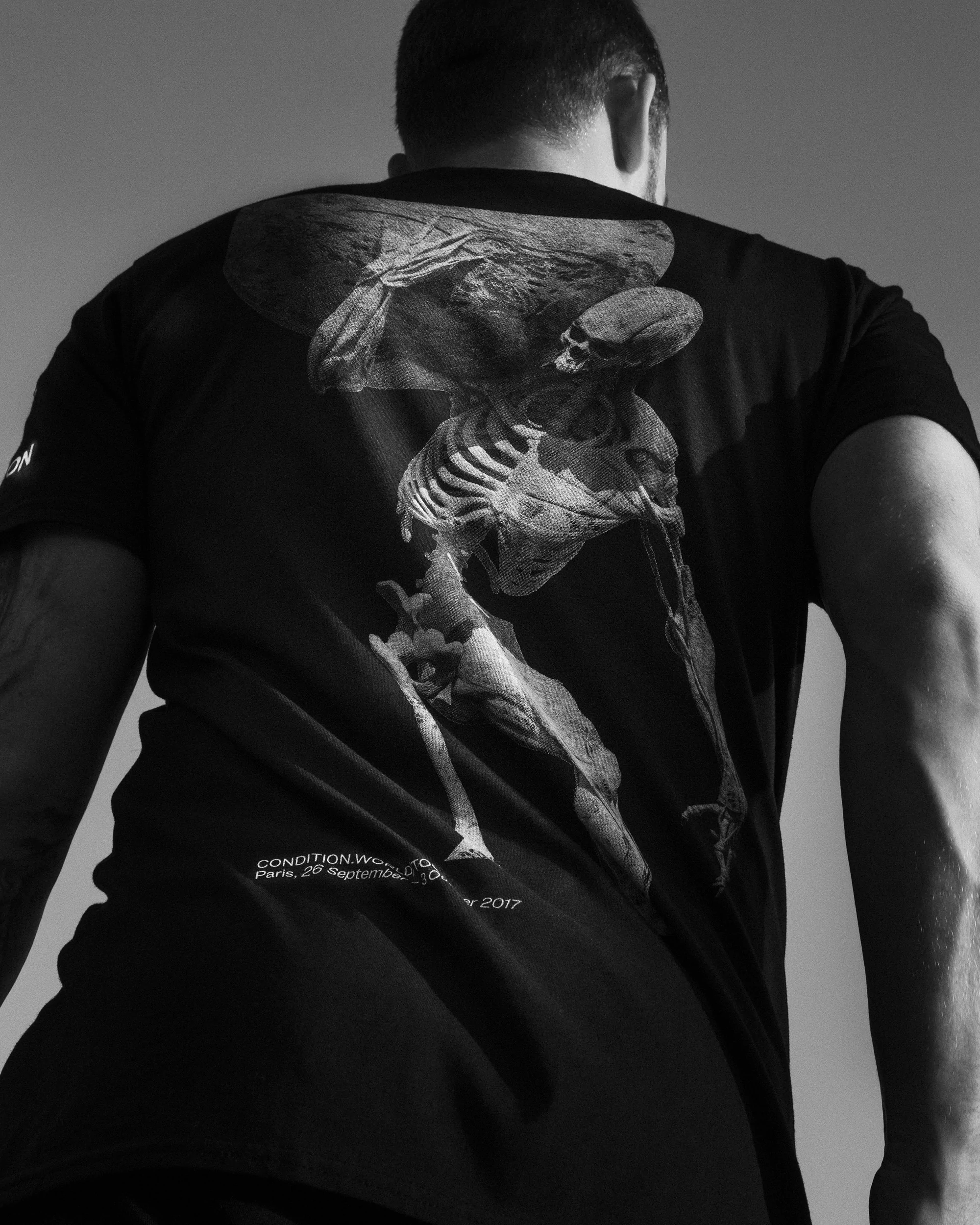 Back view of an adult male wearing a black t-shirt featuring a digital art skeleton print against a light sky background in black and white.