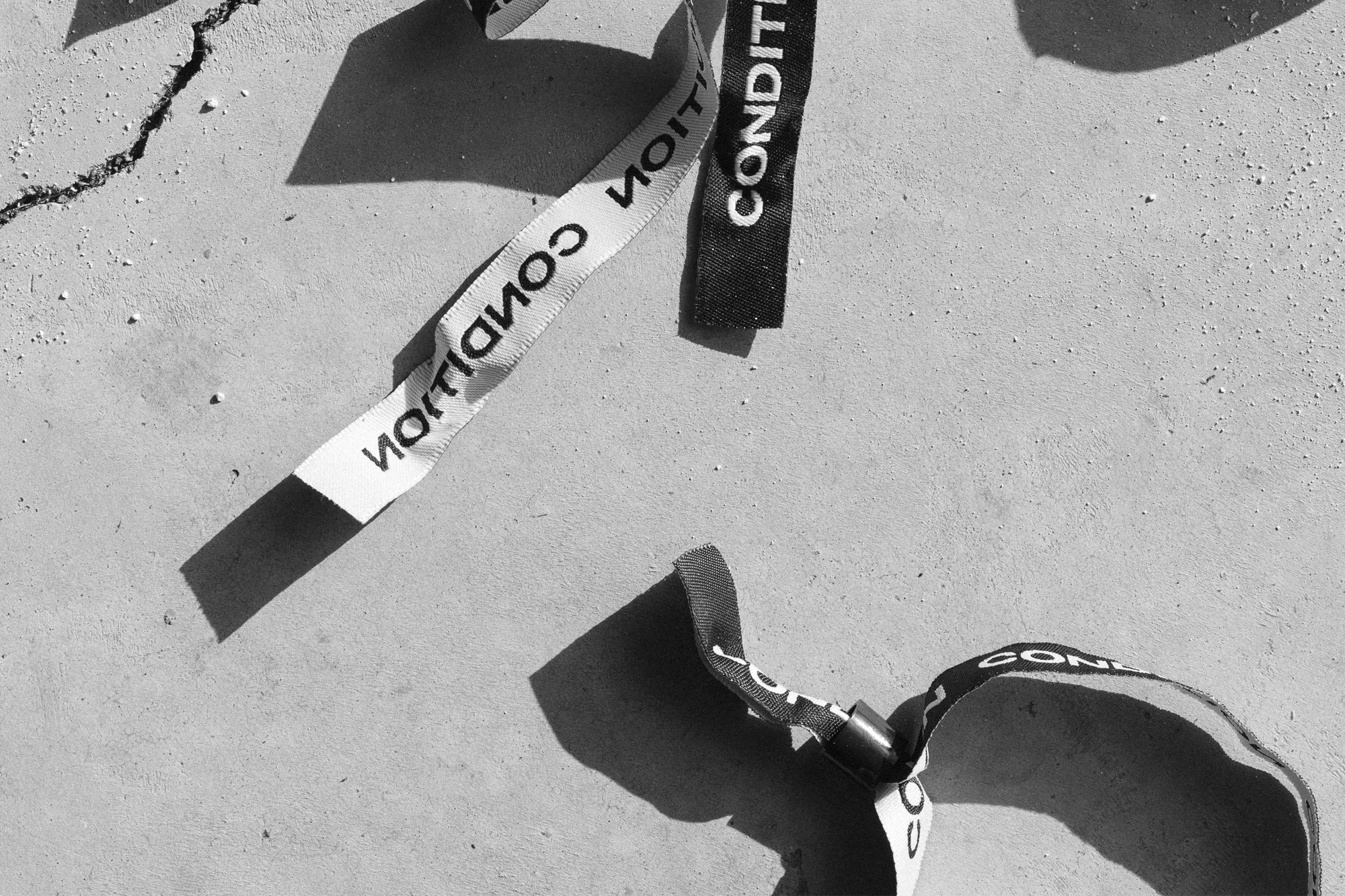 Black and white studio shot features 'Condition' branded wristbands scattered on a concrete surface with sunlight casting shadows.