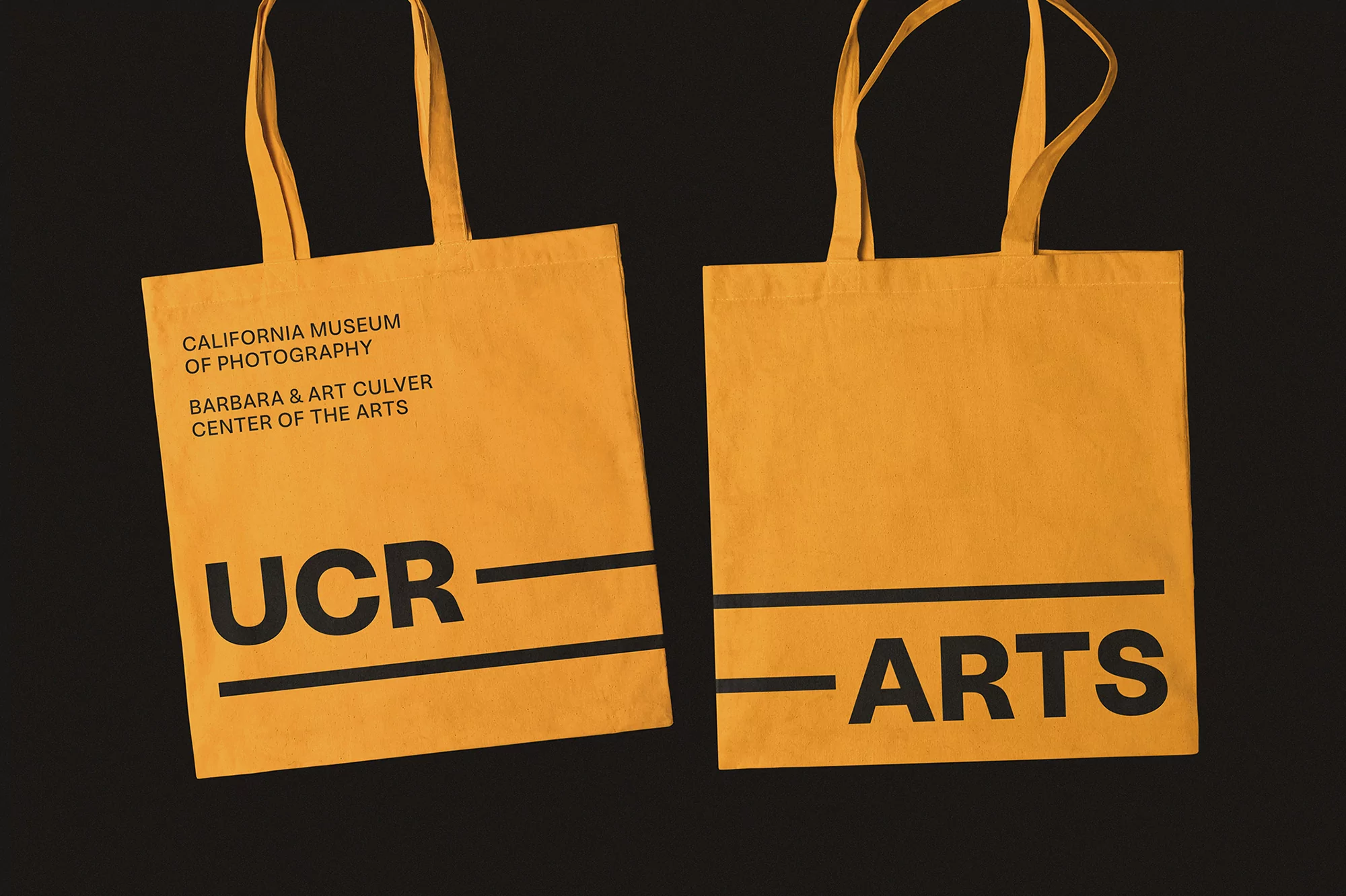 Two yellow tote bags with black text featuring 'California Museum of Photography' and 'UCR ARTS' on a dark background.