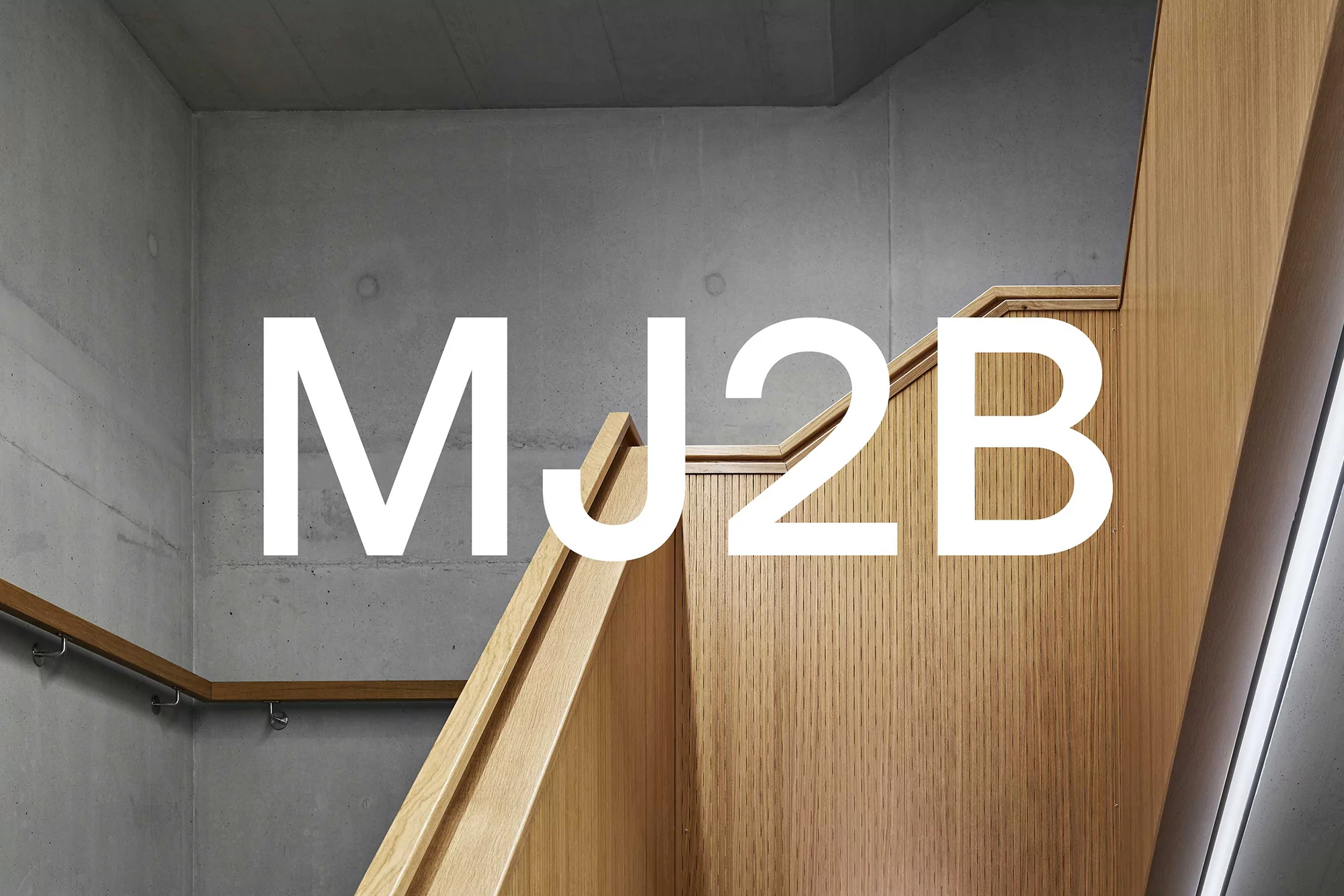 Interior view of wooden staircase with a concrete wall in the background and large white text overlay reading MJ2B.