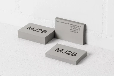 Stacks of grey business cards featuring bold black MJ2B branding resting on a textured white surface against a white wall.