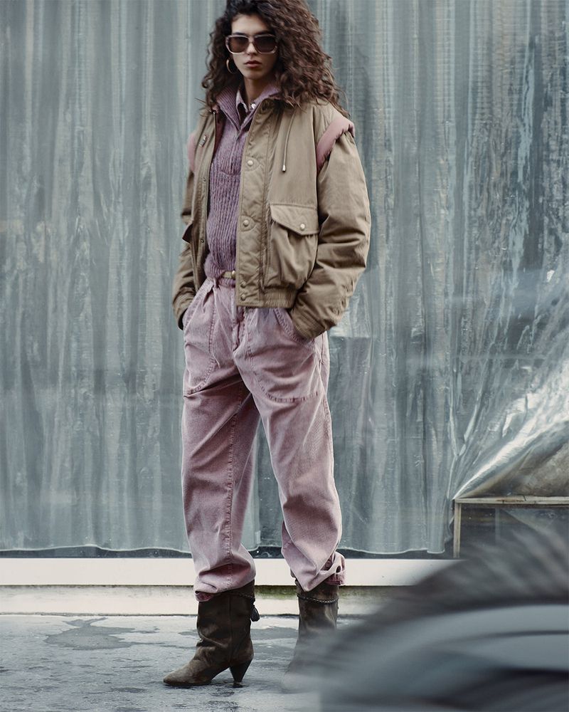 An adult female fashion model poses outdoors in a beige jacket, lavender top, pink pants, and brown heeled boots for a fashion editorial.