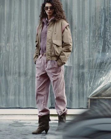 An adult female fashion model poses outdoors in a beige jacket, lavender top, pink pants, and brown heeled boots for a fashion editorial.