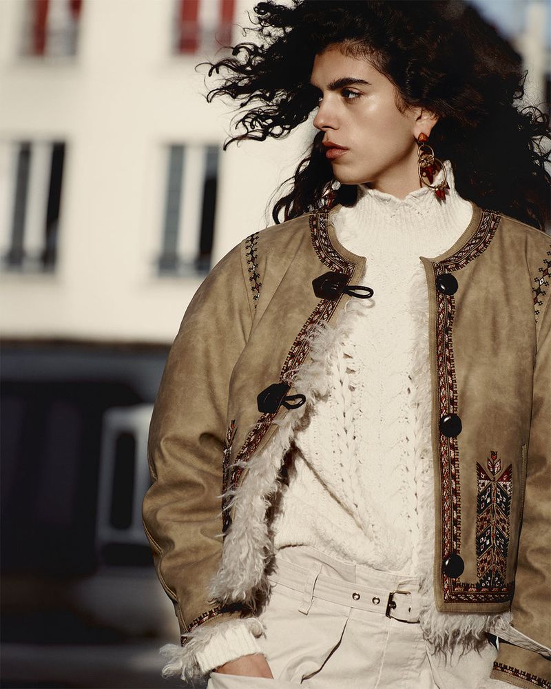 Woman with wavy hair, tan jacket, and white sweater poses in an outdoor urban setting with buildings in the soft-lit background.