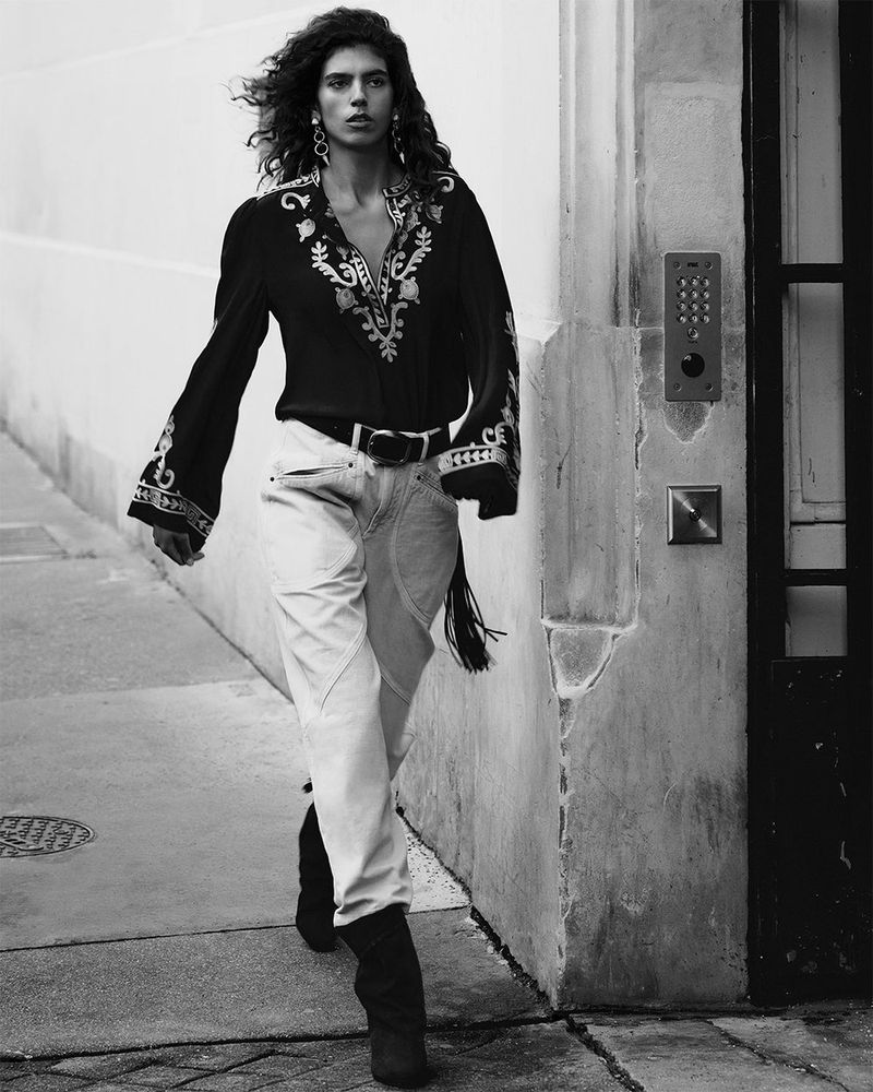 A solitary woman in a black and white fashion shot strides confidently in an urban setting wearing an embroidered top and jeans.
