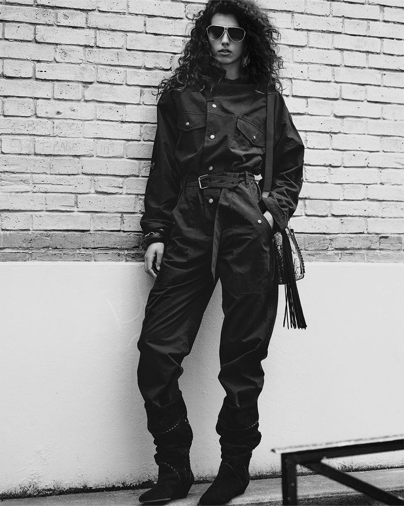 A stylish adult female poses in a black jumpsuit with a fringed bag against a brick wall, showcasing a fashion-forward urban look.