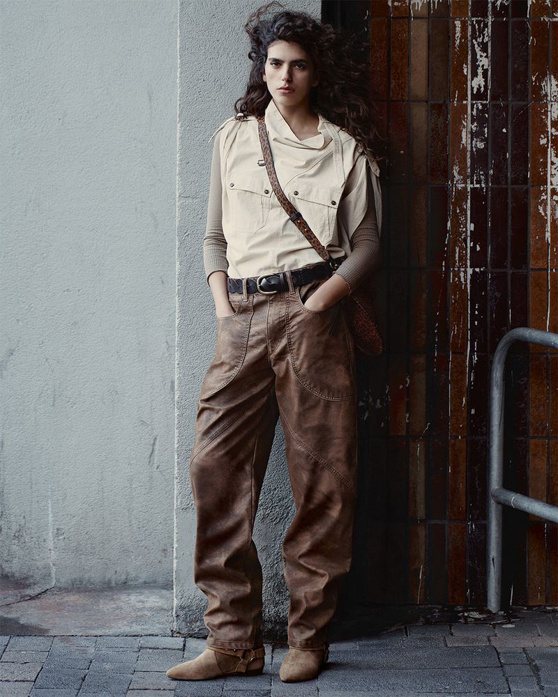 Full-shot of a young adult woman in a neutral vest, brown pants, and boots. She leans against a tiled wall on a city sidewalk.