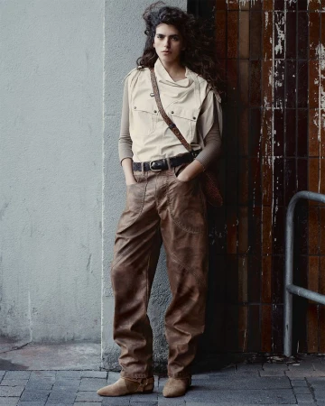 Full-shot of a young adult woman in a neutral vest, brown pants, and boots. She leans against a tiled wall on a city sidewalk.