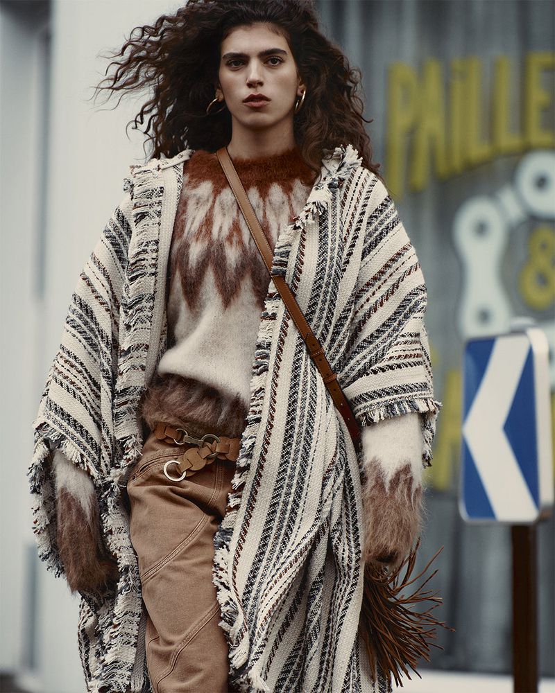 Adult woman with curly hair wears patterned sweater and poncho, accessorized with a leather belt and bag, poses outdoors in a fashion shot.