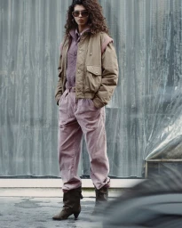 Model with long curly hair wearing sunglasses, a brown jacket, and pink trousers against a corrugated backdrop