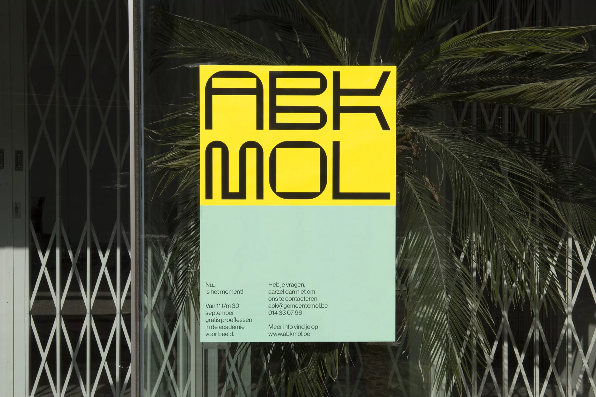 Poster with bold typography 'ABKMOL' on yellow and light green, mounted on a glass window with plant reflections and a security gate.