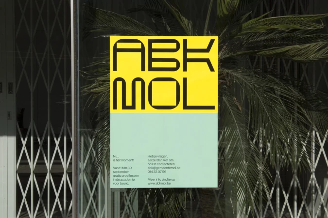 Poster with bold typography 'ABKMOL' on yellow and light green, mounted on a glass window with plant reflections and a security gate.