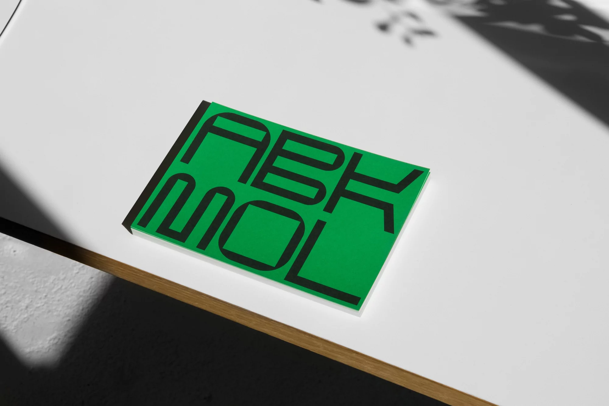 Green book with black text on a white surface, illuminated by sunlight casting shadows, creating a minimalist and graphic composition.