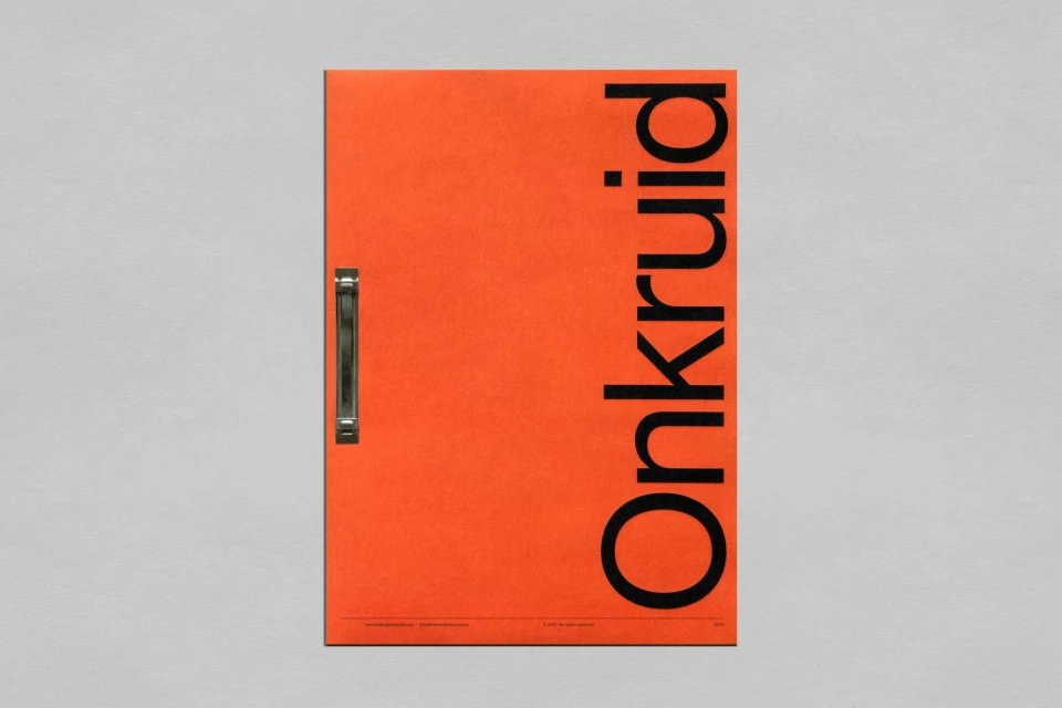 A vertically-oriented, eye-level shot of an orange booklet with a metal spine and black sans-serif typography against a white wall.