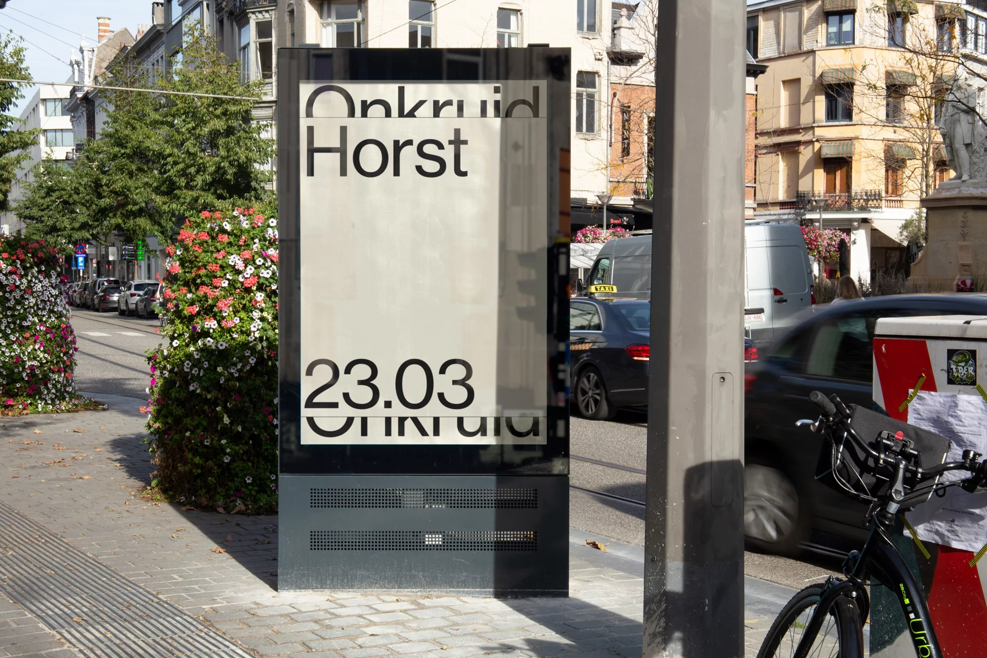 Outdoor display with 'Onkruid Horst 23.03' text on the sidewalk with trees, buildings, street traffic, and a bicycle nearby in daylight.