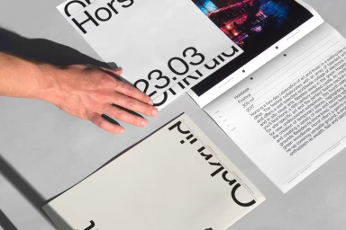Close-up of a person's hand resting on printed graphic design posters and editorial layouts featuring bold typography against a grey surface.
