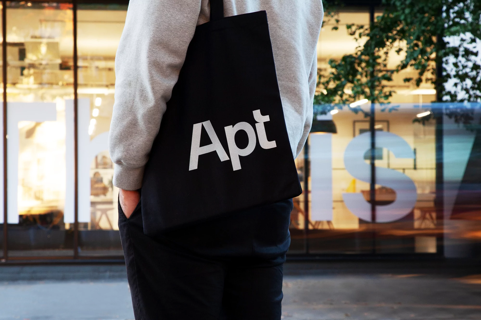 Person with a black tote bag with white 'Apt' text walking in front of a commercial storefront on an overcast urban day.