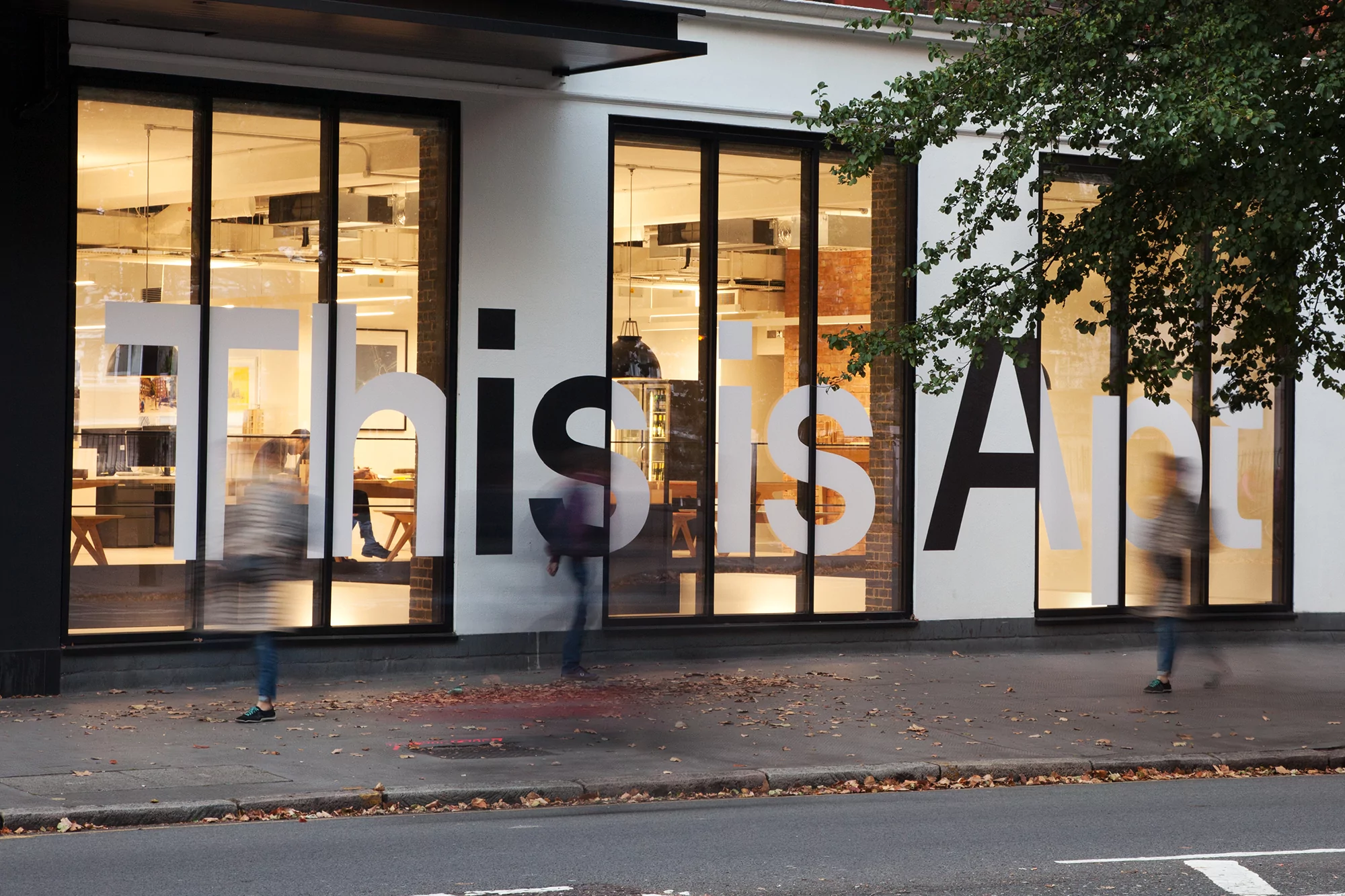 Exterior view of modern white building with large windows displaying the words 'This is Art' and blurred figures of pedestrians.