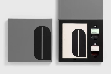 Grey presentation box containing a booklet and two glass bottles with white labels against a white background.