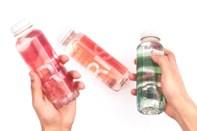 Three bottles of Burst flavored water in red, orange, and green colors are held by two hands against a plain white backdrop.