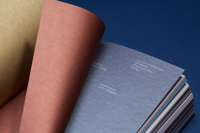 Close-up of a paper sample book with multiple textured sheets in different colors like yellow, coral, and blue on a blue background.