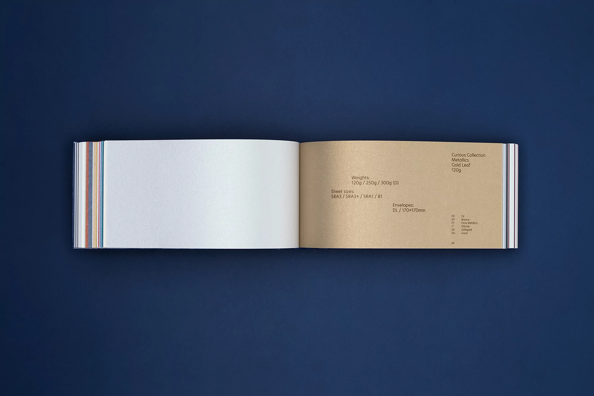 Open paper sample book showcasing textured white and gold leaf paper against a dark blue background, ideal for design and material selection.