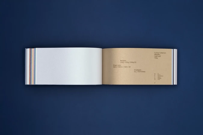 Open paper sample book showcasing textured white and gold leaf paper against a dark blue background, ideal for design and material selection.
