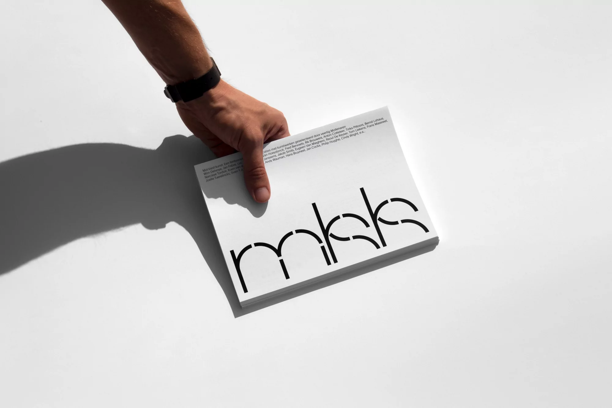 Hand holds a white book with bold black lettering, photographed in a brightly lit studio, creating sharp shadows and a clean aesthetic.