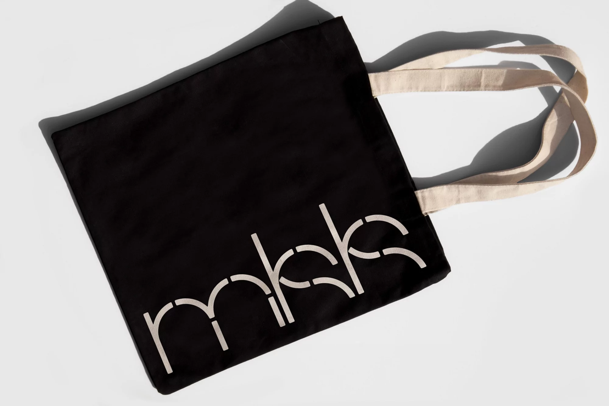 Black tote bag with white stencil-style lettering on a white background, simple studio product shot with natural light and shadow.