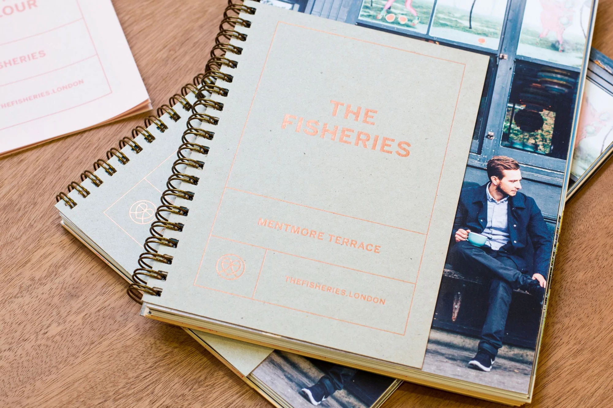Close-up of 'The Fisheries' notebooks on a wooden table, featuring a man with a coffee on one cover and branding on the other.