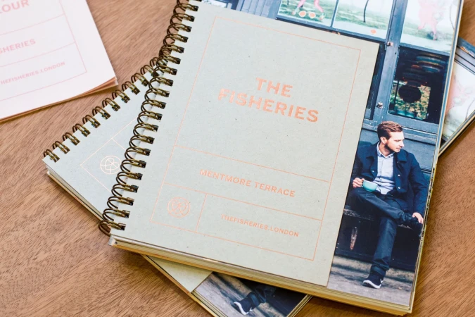 Close-up of 'The Fisheries' notebooks on a wooden table, featuring a man with a coffee on one cover and branding on the other.