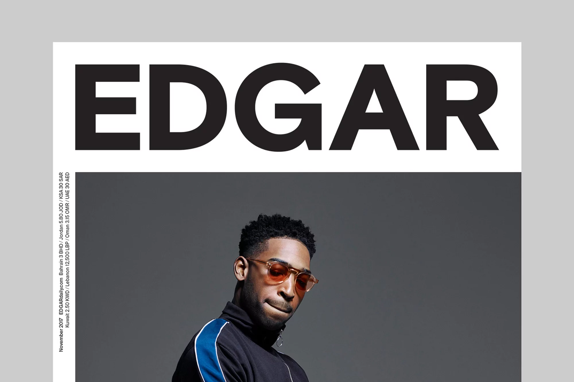 Magazine cover featuring a stylish man with tan sunglasses, wearing a black tracksuit with blue stripes, against a grey backdrop.
