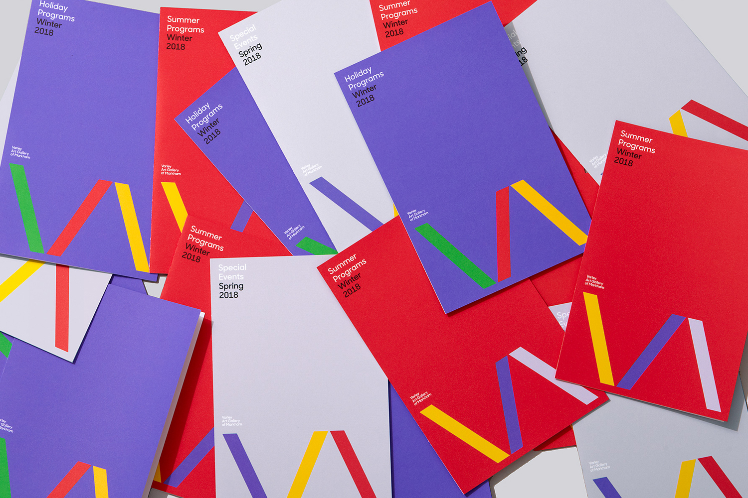 Multiple red, purple, and grey program brochures with colorful geometric designs, titled Winter and Spring Programs 2018, on a flat surface.