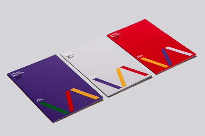 Three vividly colored program brochures, titled Holiday Programs Winter 2018, Special Events Spring 2018, and Summer Programs 2018, displayed on gray.