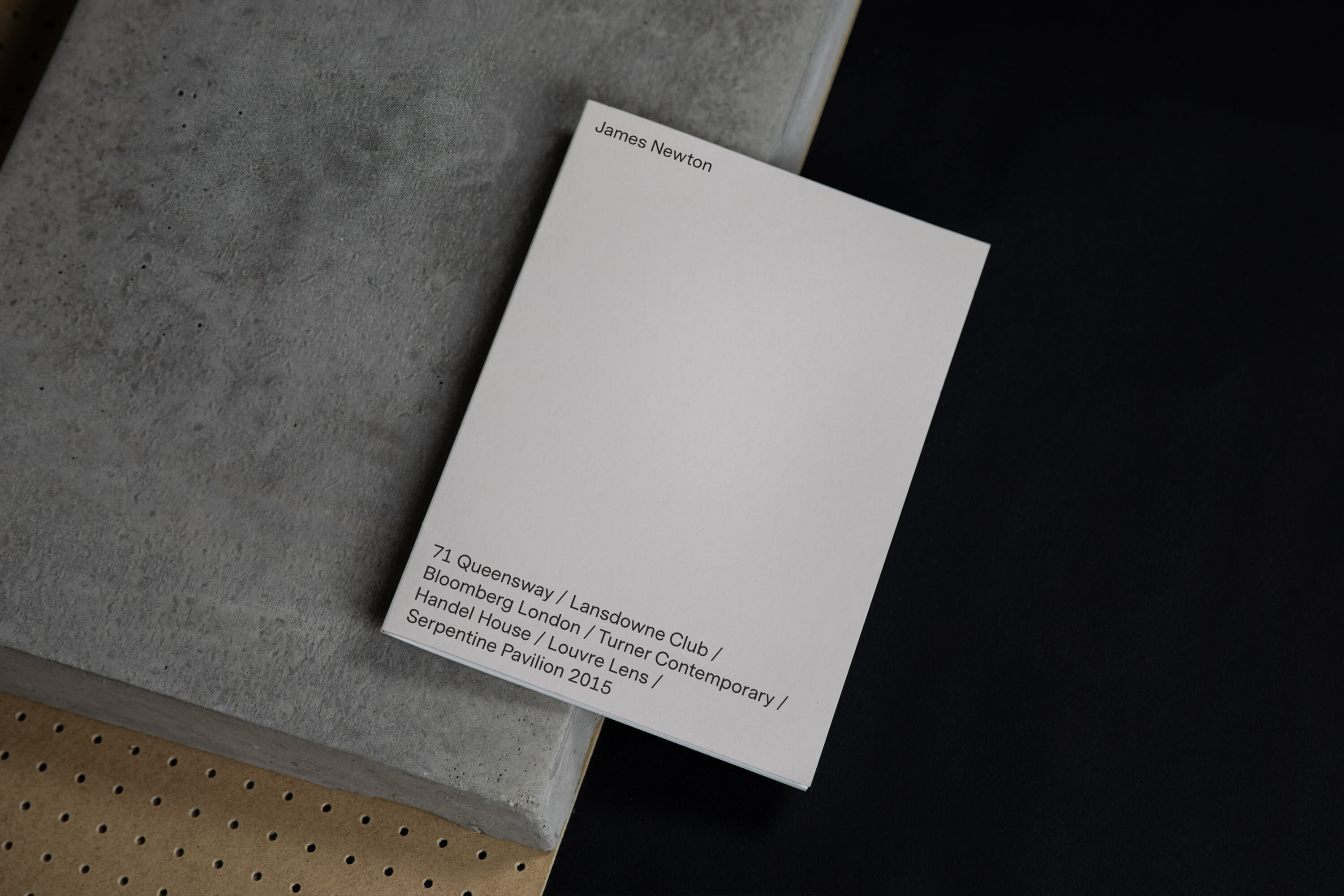 White rectangular card featuring black text 'James Newton' on a gray concrete slab against a dark backdrop in a neutral studio setting.