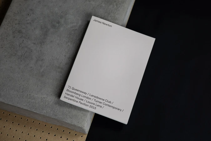 White rectangular card featuring black text 'James Newton' on a gray concrete slab against a dark backdrop in a neutral studio setting.
