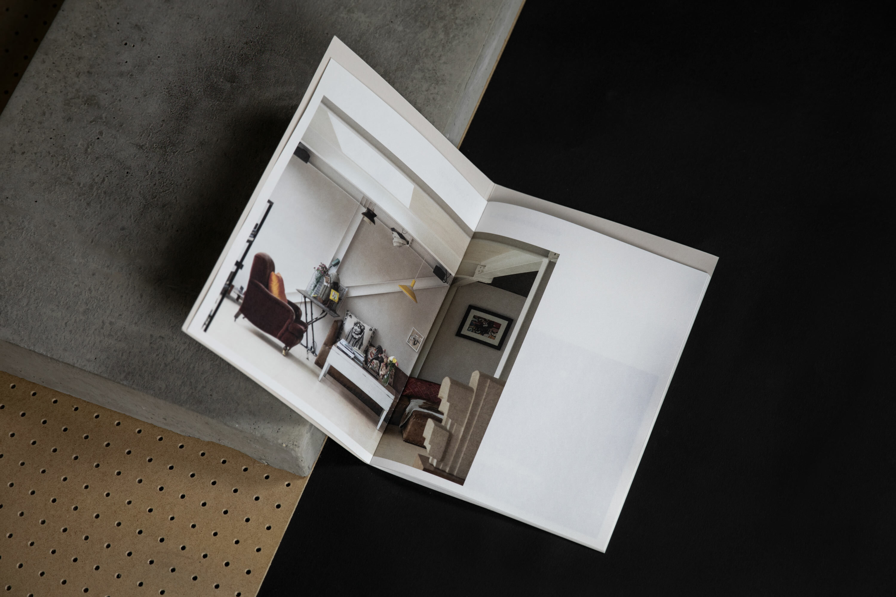 Open photo book on concrete, featuring an interior design image with a chair, table, decor against a white-walled room with a fan.