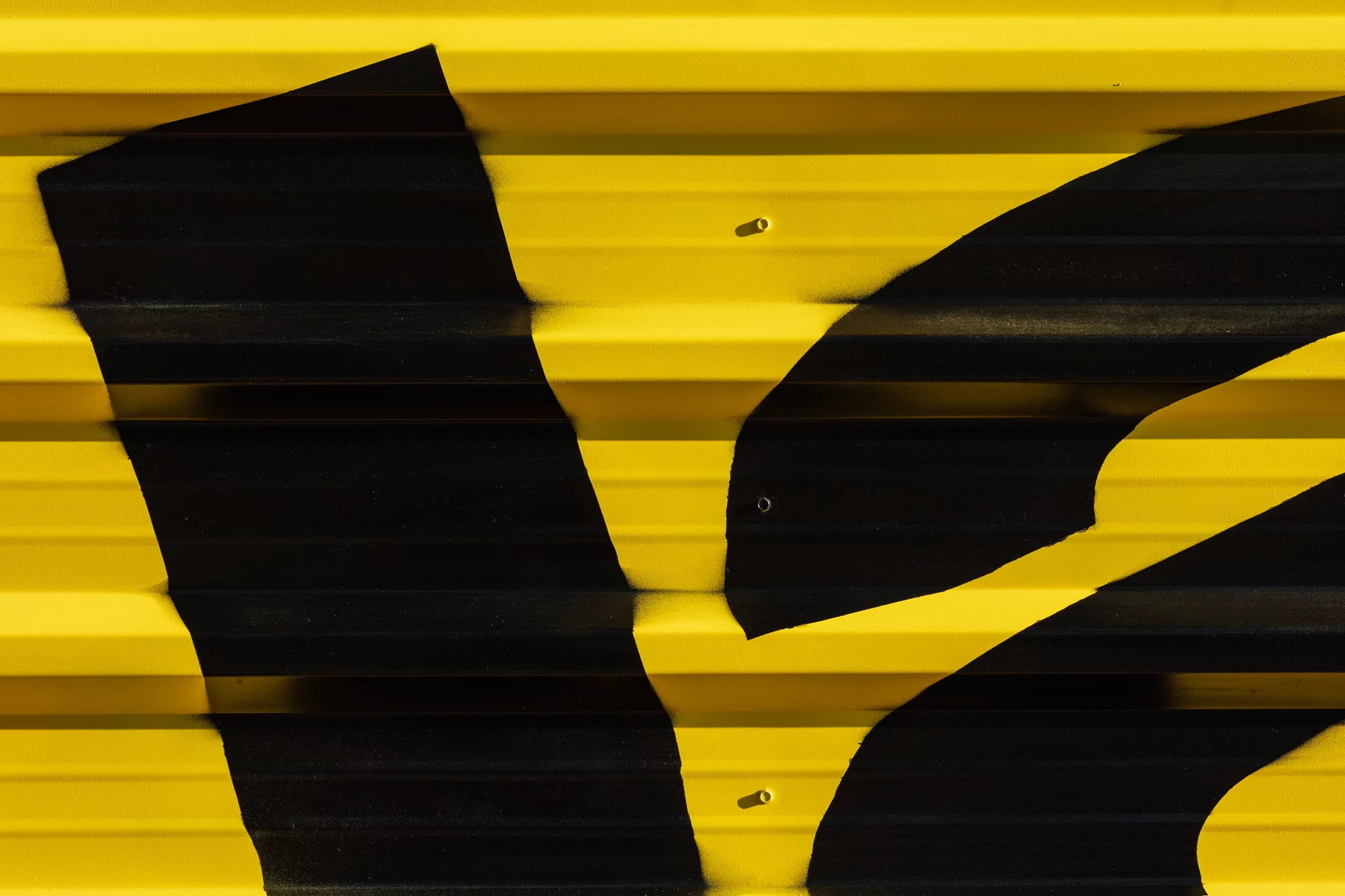 Abstract close-up of black painted lines against a yellow ribbed surface featuring an industrial metal wall with visible fasteners.