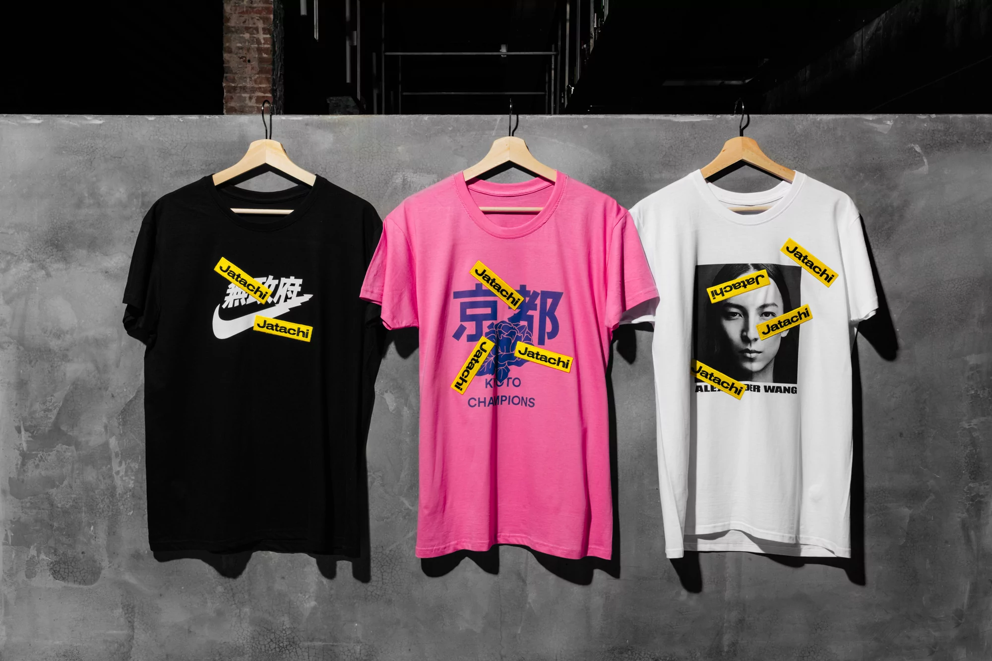 Three graphic t-shirts hang on wooden hangers against a gray wall, featuring designs with logos and Asian characters.