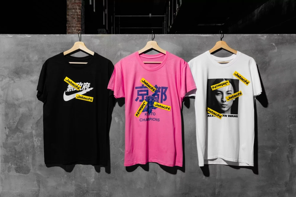 Three graphic t-shirts hang on wooden hangers against a gray wall, featuring designs with logos and Asian characters.