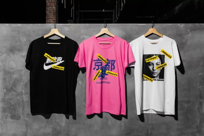 Three graphic t-shirts hang on wooden hangers against a gray wall, featuring designs with logos and Asian characters.