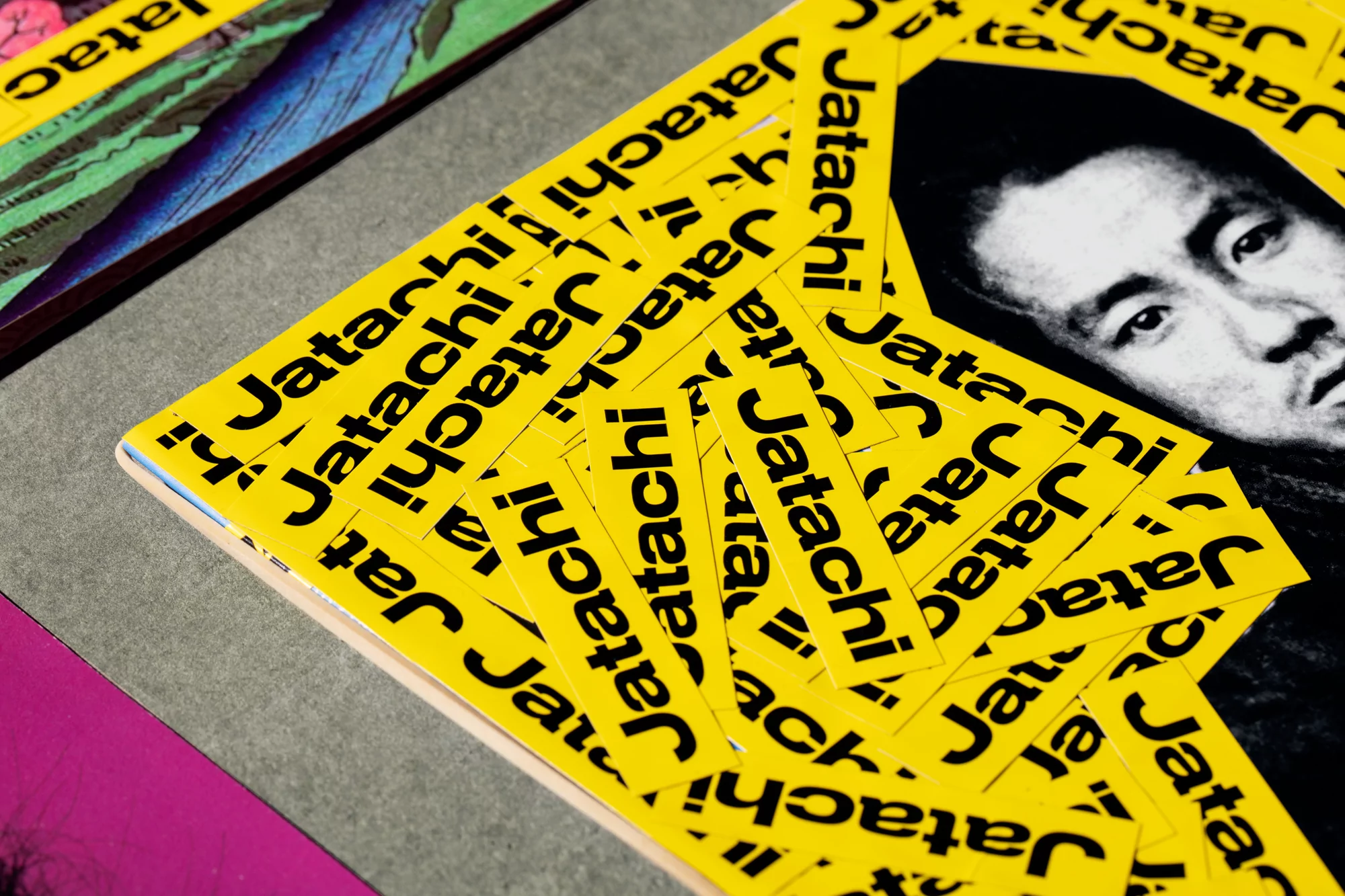 Close-up photograph of yellow Jatachi branded stickers stacked on top of a book with a portrait on the cover, gray and pink background.