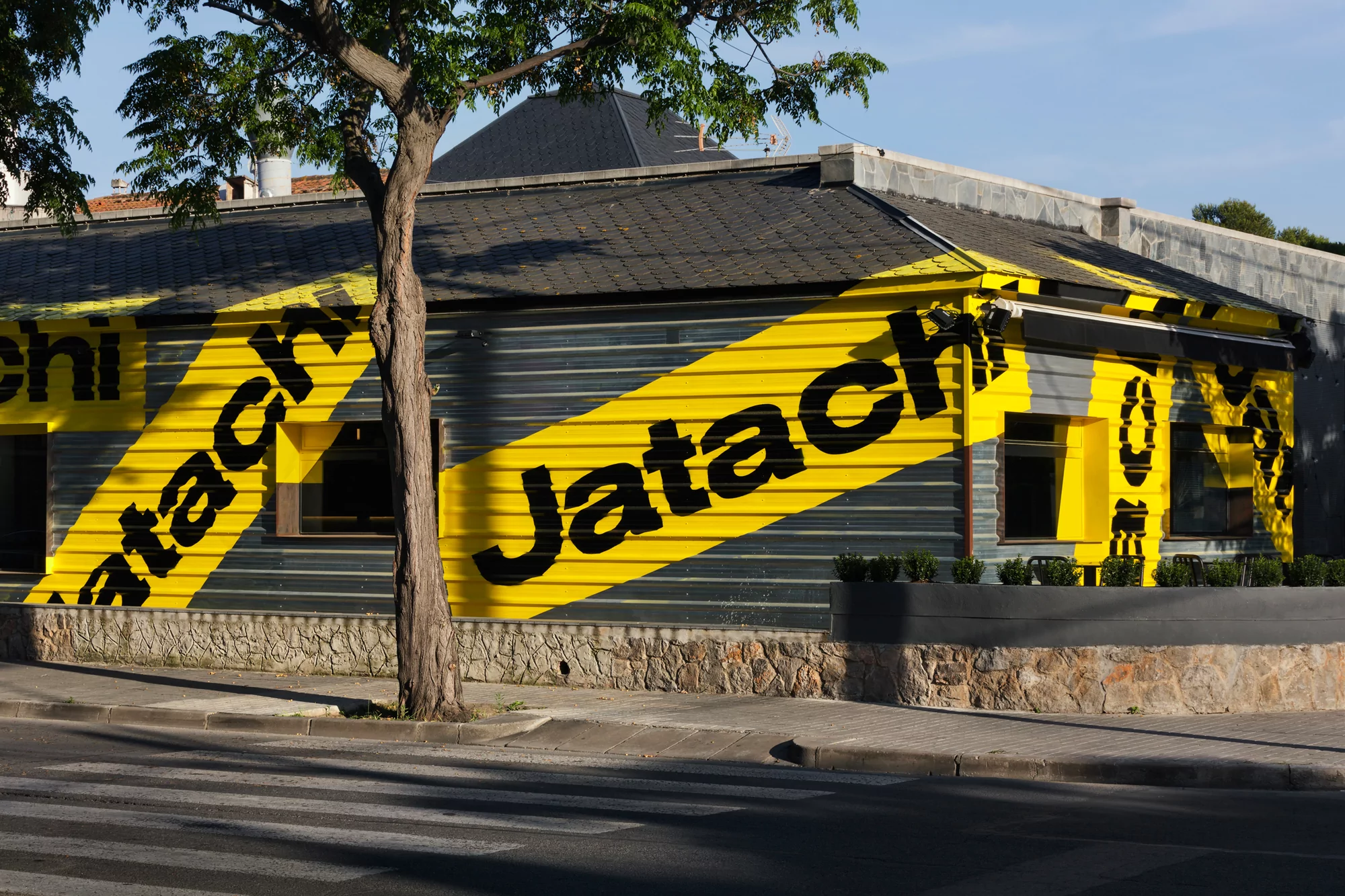 Exterior shot of a commercial building featuring a facade with a gray corrugated metal and stone base, marked with bold yellow and black stripes.
