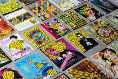 Overhead view of multiple magazines and publications arranged on a concrete surface, covered with yellow Jatachi caution tape stickers.