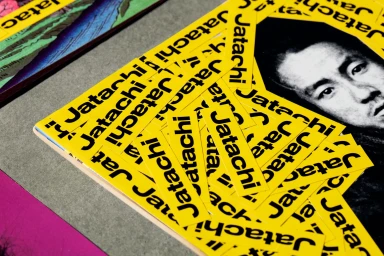 Close-up of bright yellow Jatachi brand stickers scattered across a grey textured surface next to printed media.