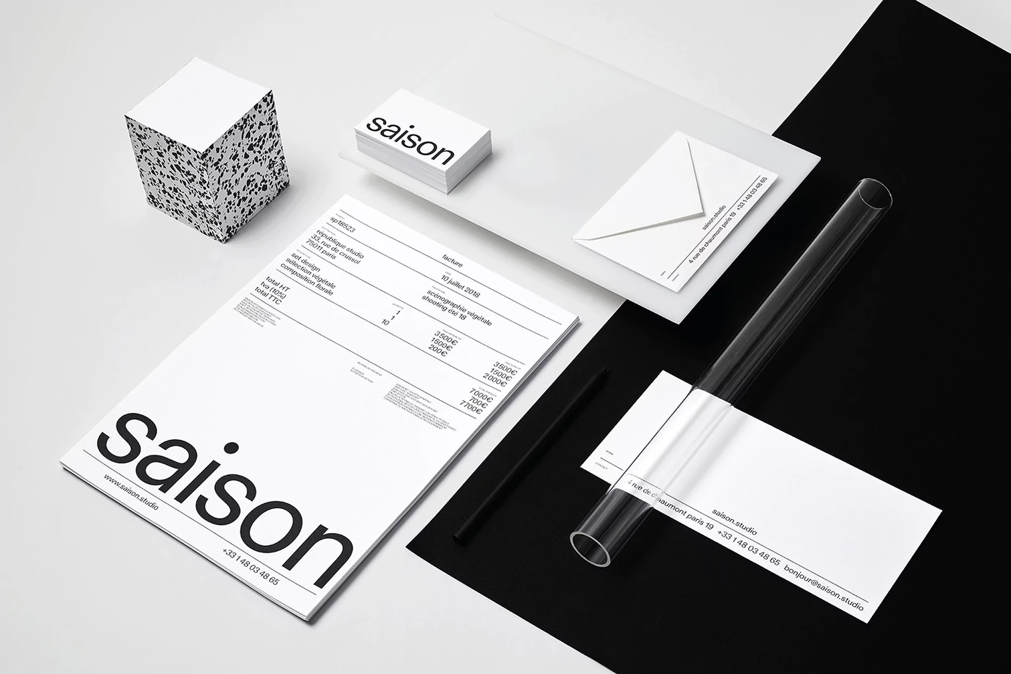 Minimalist flat lay composition of stationery including a cube, business cards, letter, invoice, pen, and cylinder, on a contrasting black and white surface.