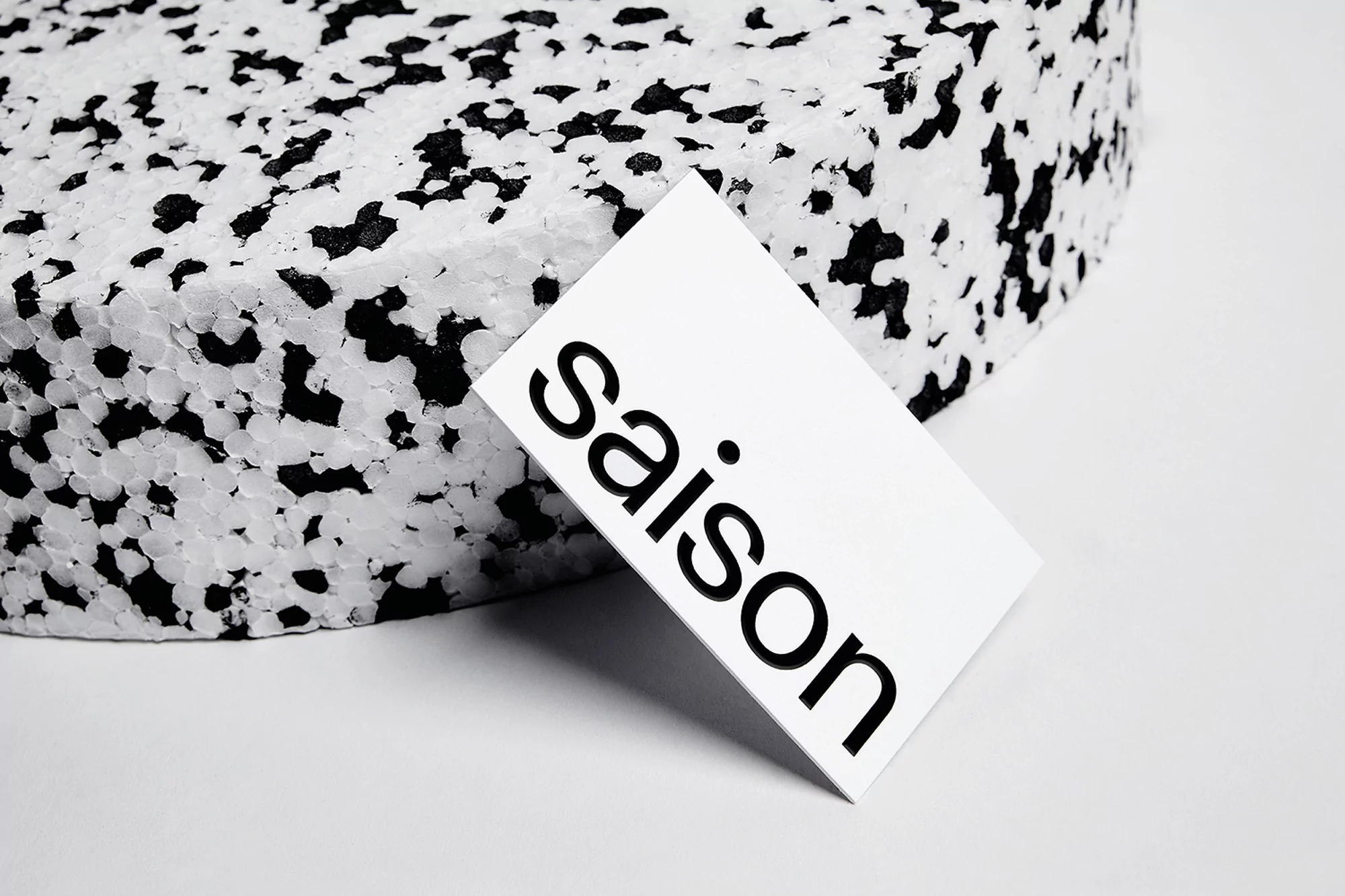 A white card with the word 'saison' in black lettering leans against a textured white and black object in a minimalist setting.