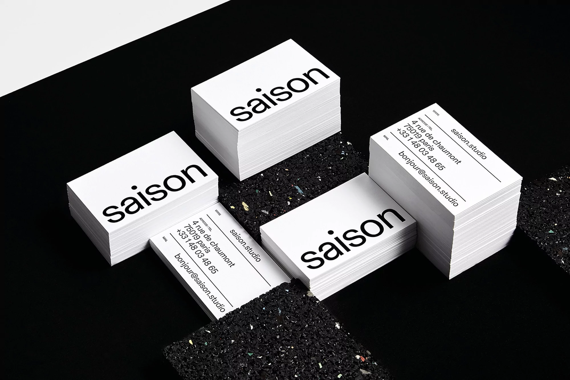 Stacks of minimalist business cards on a black surface, featuring the word 'saison' and contact information for saison.studio in Paris.